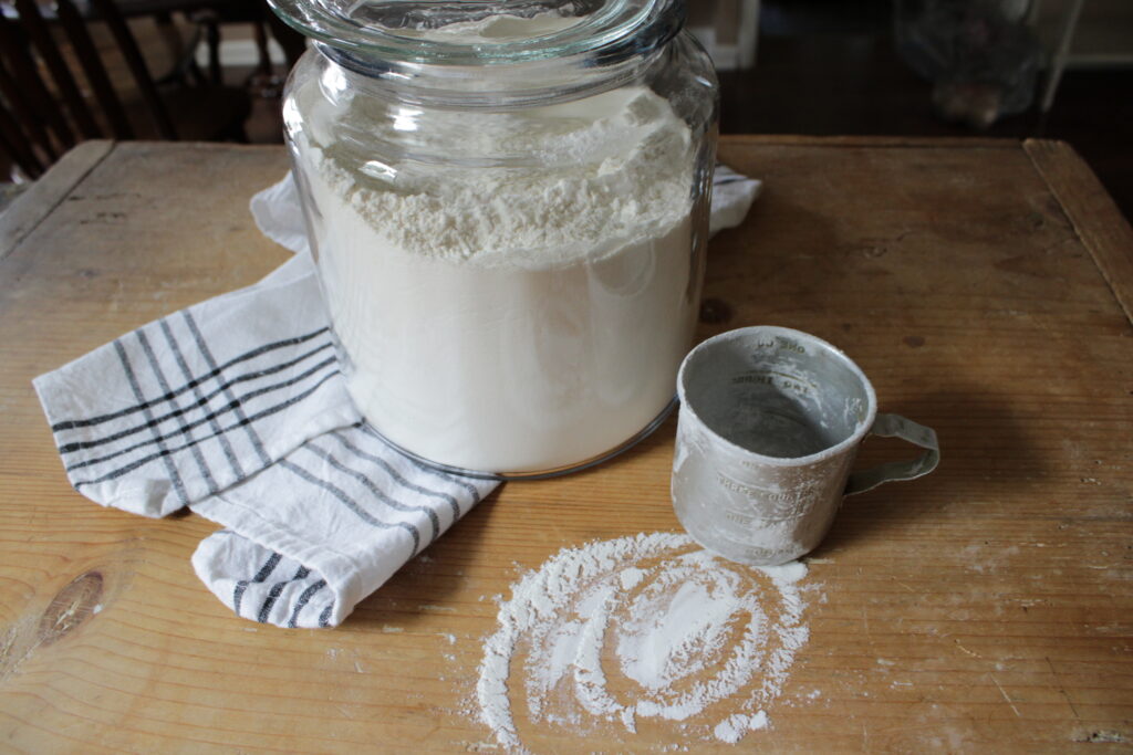 Glass jar of all purpose flour with measuring cup sitting on wooden cutting board surrounded by spilled flour.